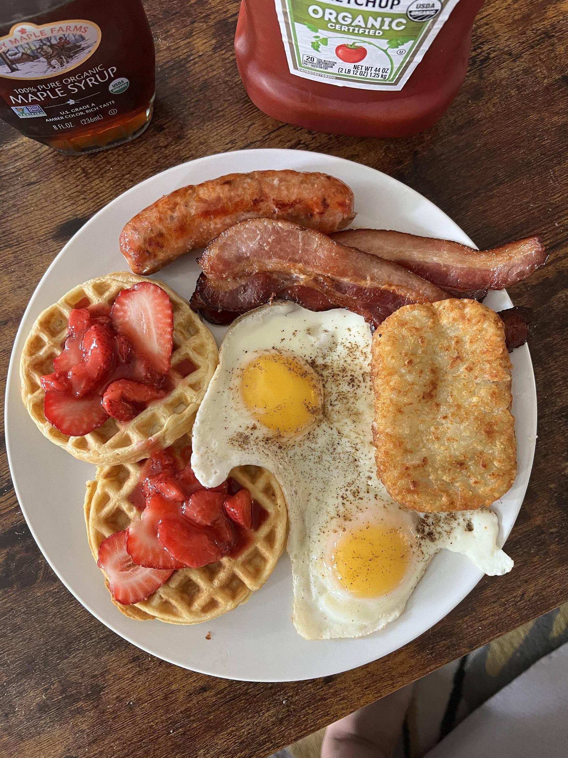 A breakfast plate with two waffles topped with strawberries, two sunny side up eggs, a hash brown, bacon, and sausage. Bottles of maple syrup and organic ketchup are in the background.