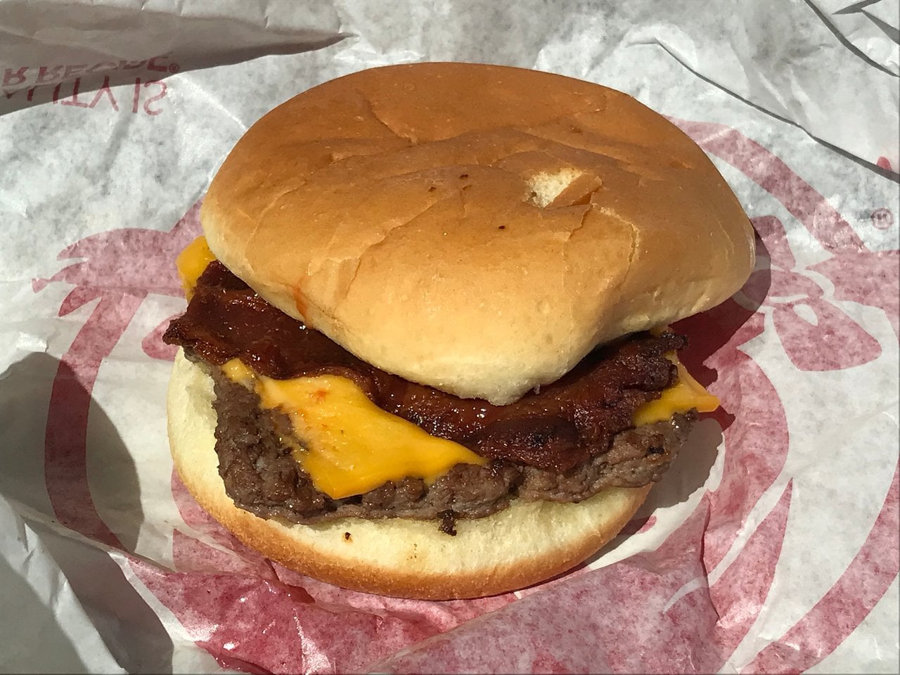 A cheeseburger with a beef patty, cheddar cheese, and bacon inside a sandwich bun, placed on crumpled fast-food wrapper paper.