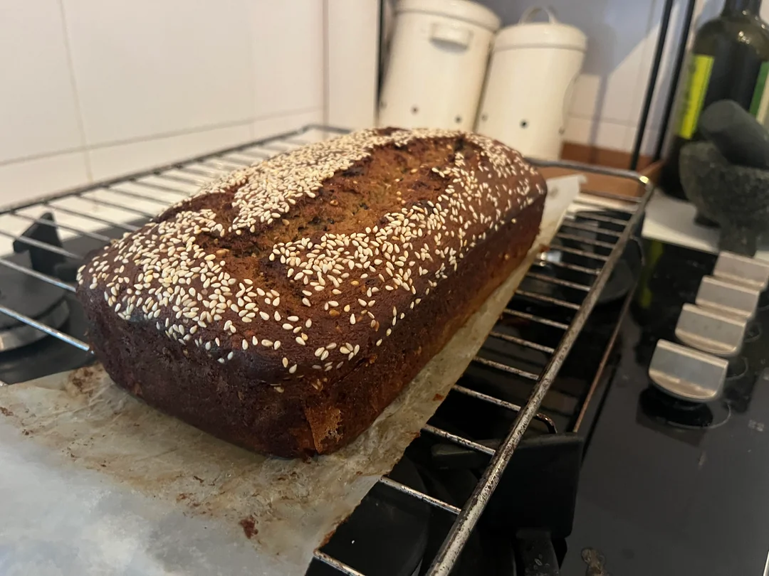 A loaf of homemade bread topped with sesame seeds cools on a wire rack, with parchment paper underneath. The bread is golden brown, and kitchen canisters and a stove are visible in the background.