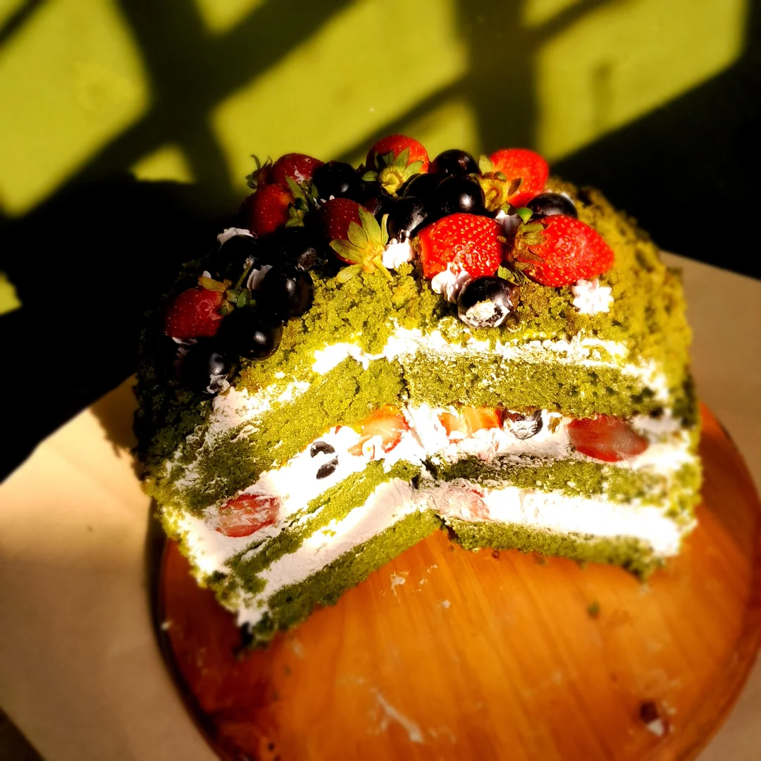 A sliced matcha cake with layers of cream and berries, topped with strawberries and blueberries, sits on a wooden board in sunlight.