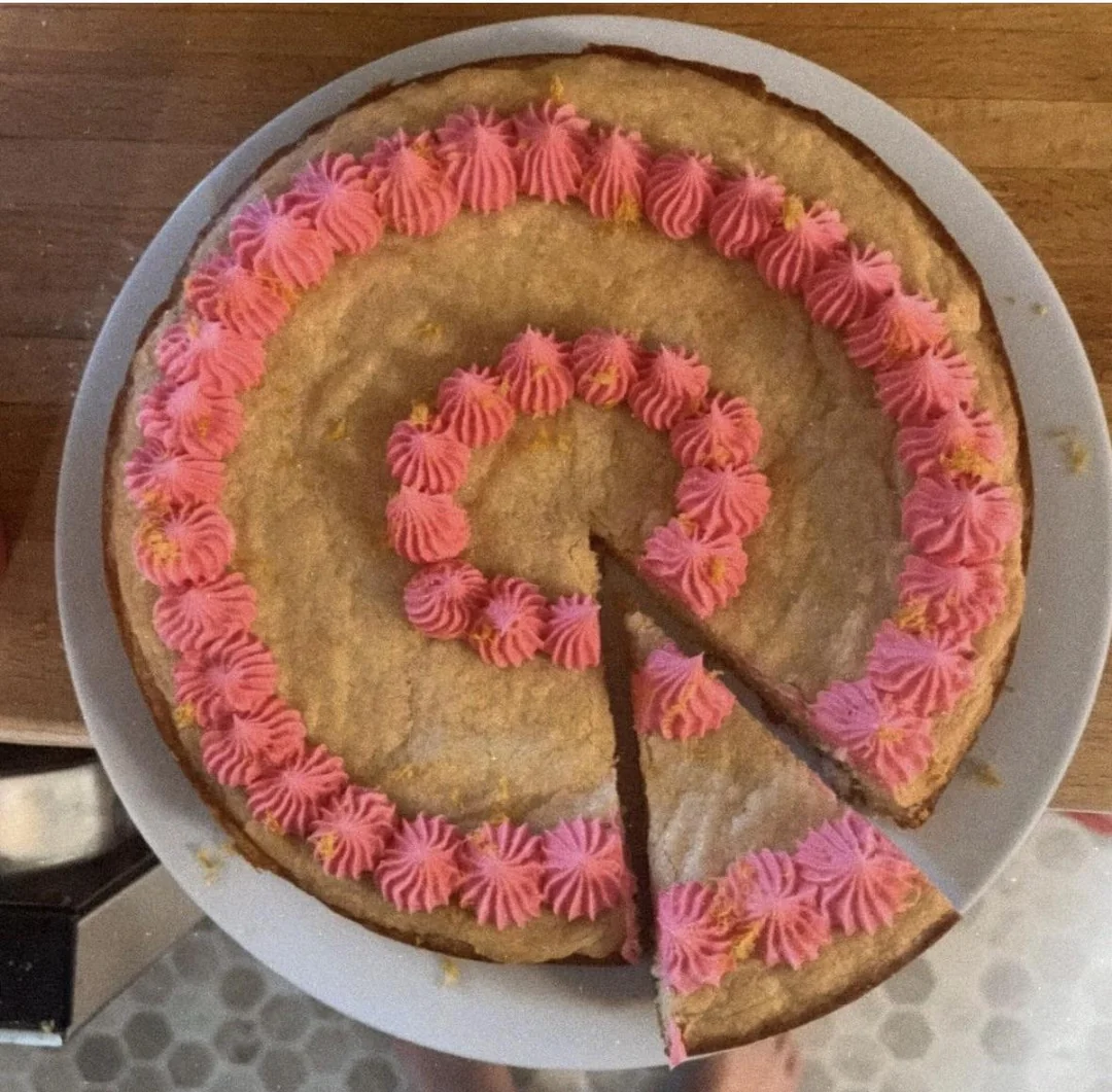 A round cookie cake on a white plate, topped with pink frosting in a spiral pattern. One triangular slice has been cut and slightly pulled out from the whole cake.