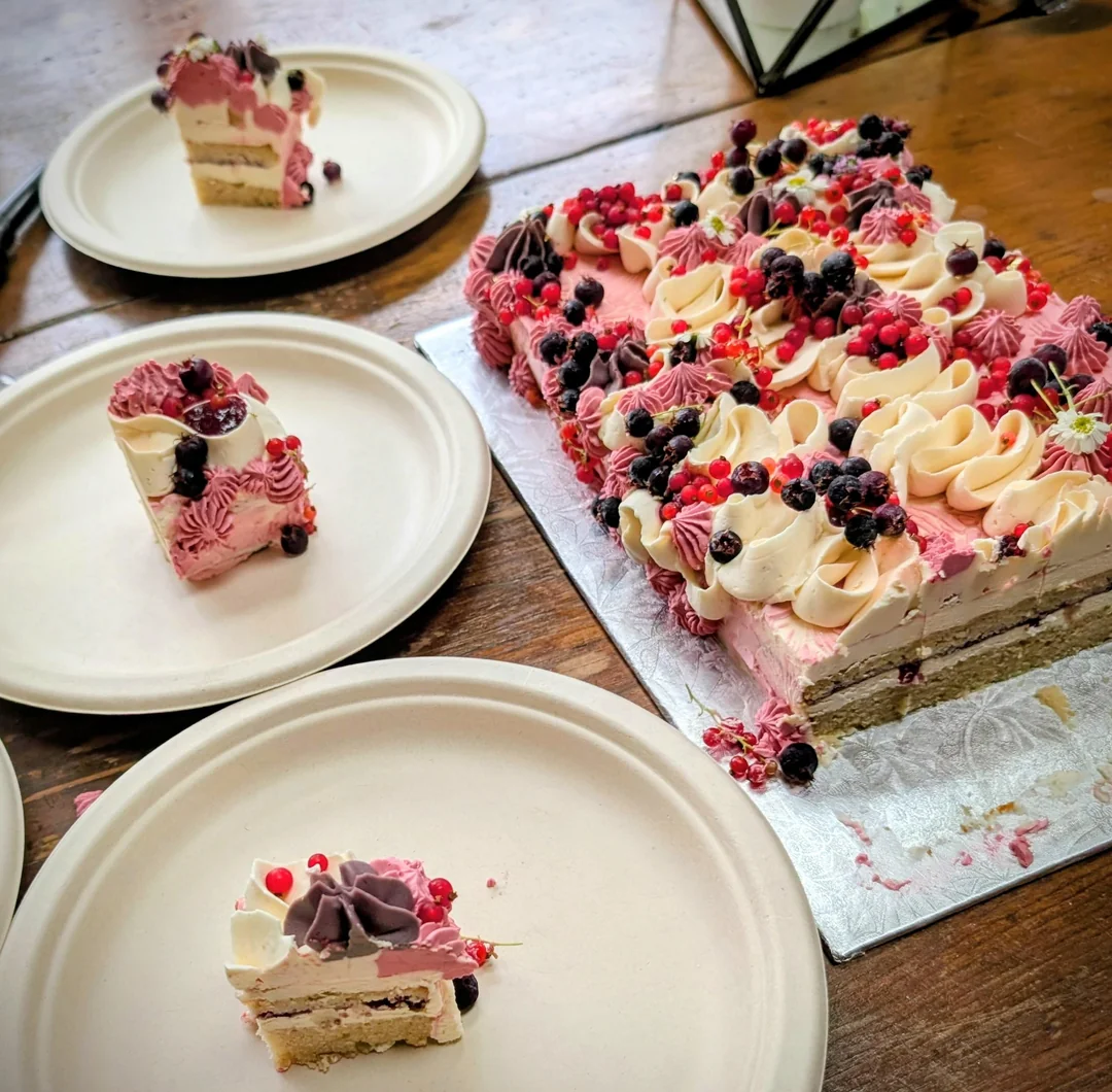 Rectangular berry-topped cake on a tray with three plates, each holding a slice. The cake has pink and cream frosting, berries, and sponge layers, all set on a wooden table.