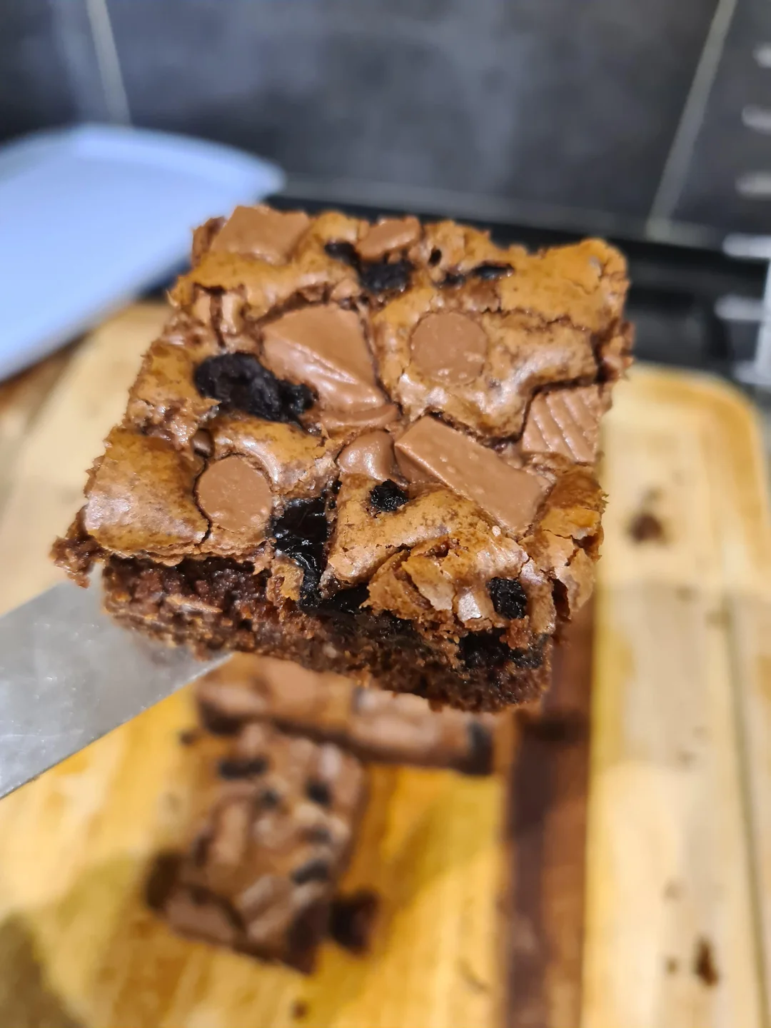 A close-up of a chocolate brownie topped with chocolate chunks and chips, held on a spatula above a wooden cutting board.