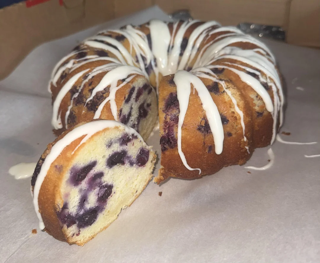A blueberry bundt cake with white icing drizzled on top sits on parchment paper. One slice is cut and slightly pulled out, revealing blueberries inside the moist cake.