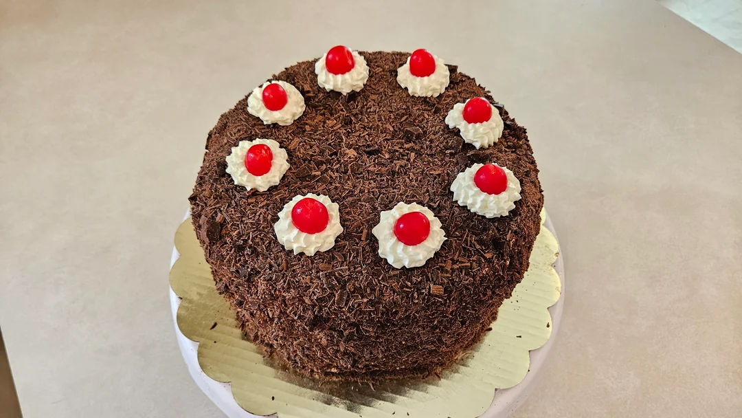 A round chocolate cake topped with eight whipped cream dollops, each decorated with a red cherry, sits on a gold scalloped cake board.