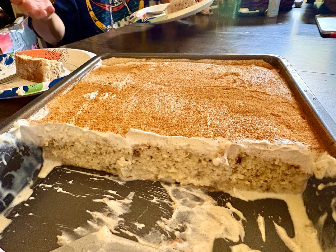 A large rectangular cake with white frosting and cinnamon topping sits in a baking pan with several pieces cut out. A person in the background holds a plate with a slice of the cake.