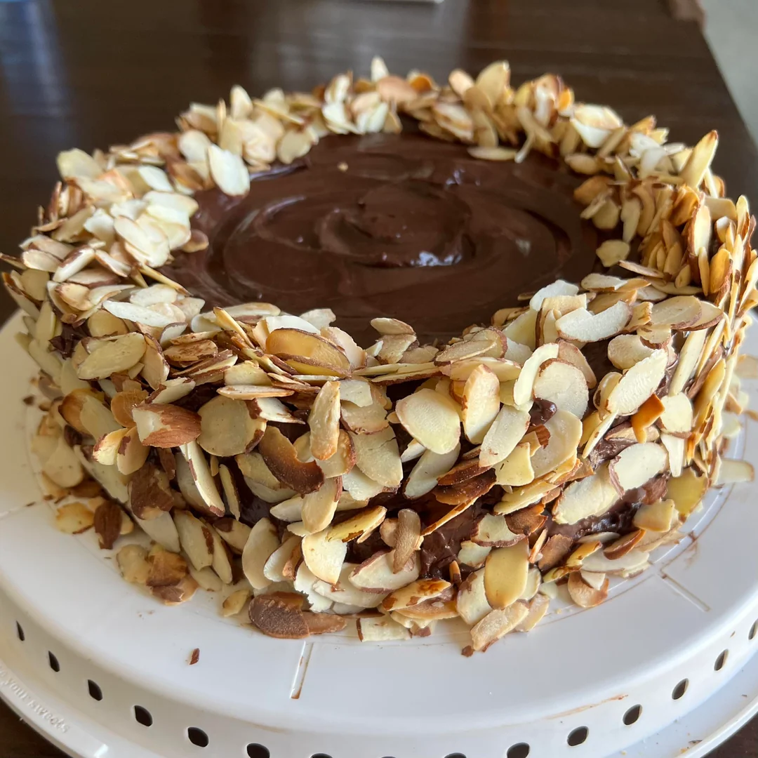 A round chocolate cake covered with sliced almonds on the sides and topped with glossy chocolate frosting, sitting on a white cake stand.