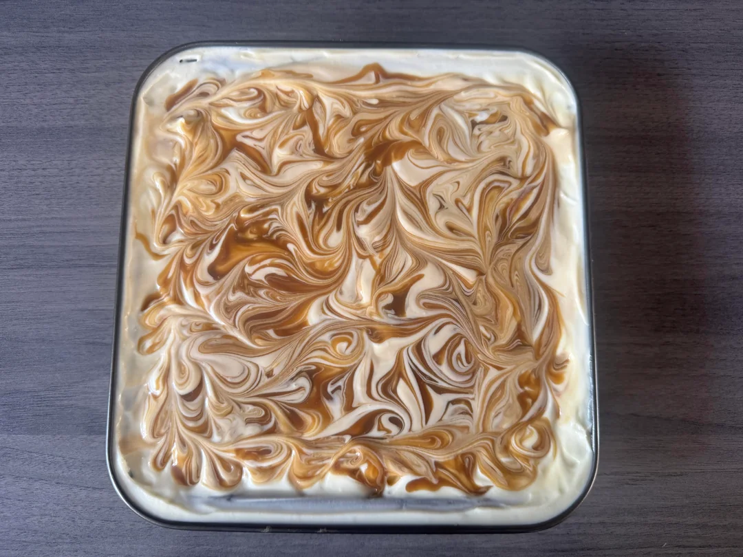A square glass dish filled with creamy dessert, topped with swirled caramel and white cream, creating a marbled pattern, placed on a dark wooden surface.