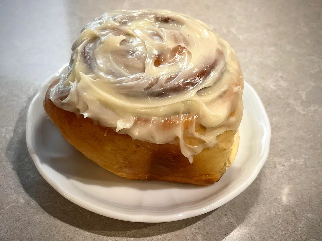 A cinnamon roll topped with creamy white icing sits on a small white plate on a light-colored countertop.