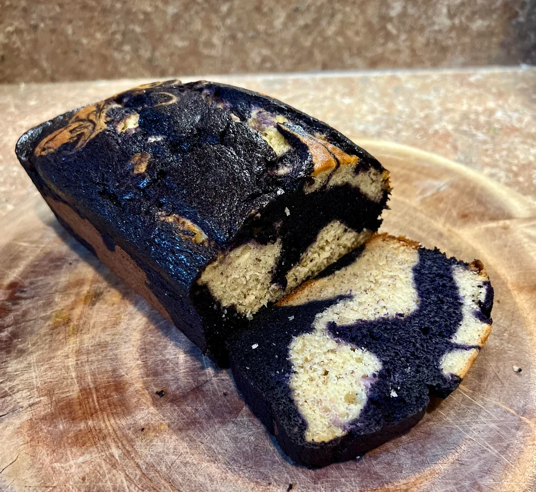 A sliced loaf of marble cake with swirls of dark chocolate and light vanilla on a round wooden cutting board, set against a brown speckled countertop.