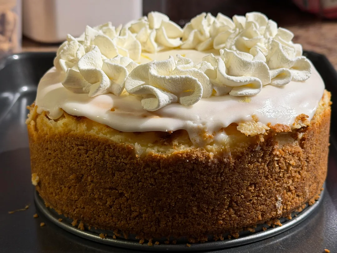 A cheesecake with a thick graham cracker crust, topped with white icing and decorative swirls of whipped cream, sits on a metal baking pan.