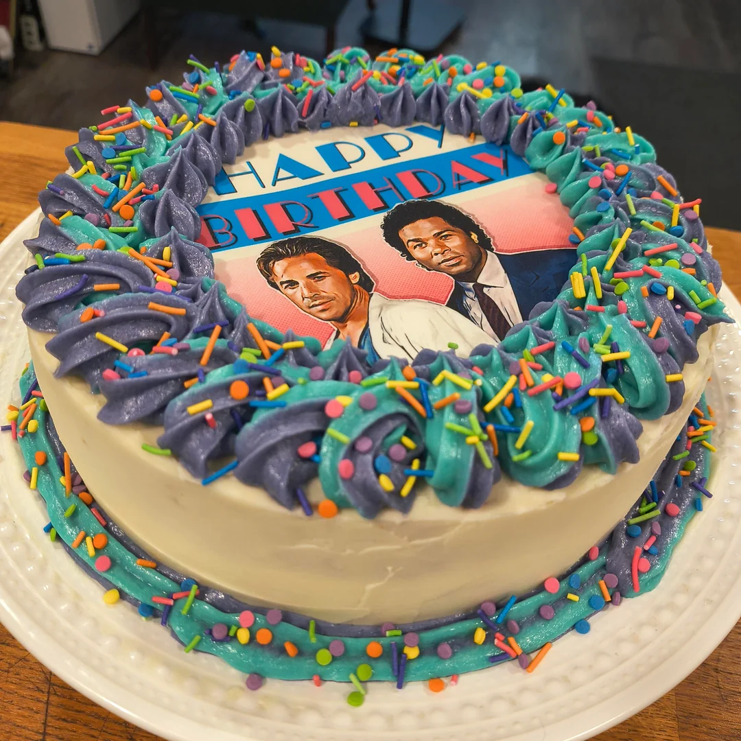 A birthday cake with purple and teal frosting, colorful sprinkles, and an edible image of two men in suits with “Happy Birthday” written above them. The cake sits on a white plate.