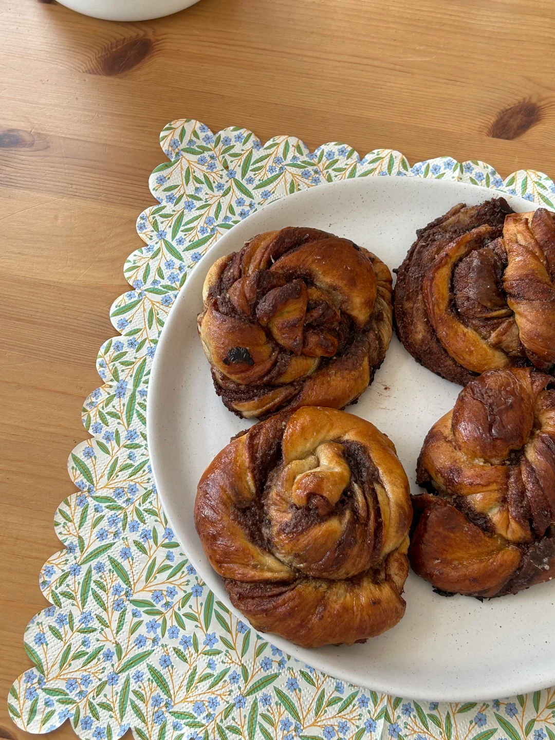 Homemade baked goods. A white plate with four cinnamon buns sits on a wooden table, atop a floral-patterned placemat with scalloped edges.