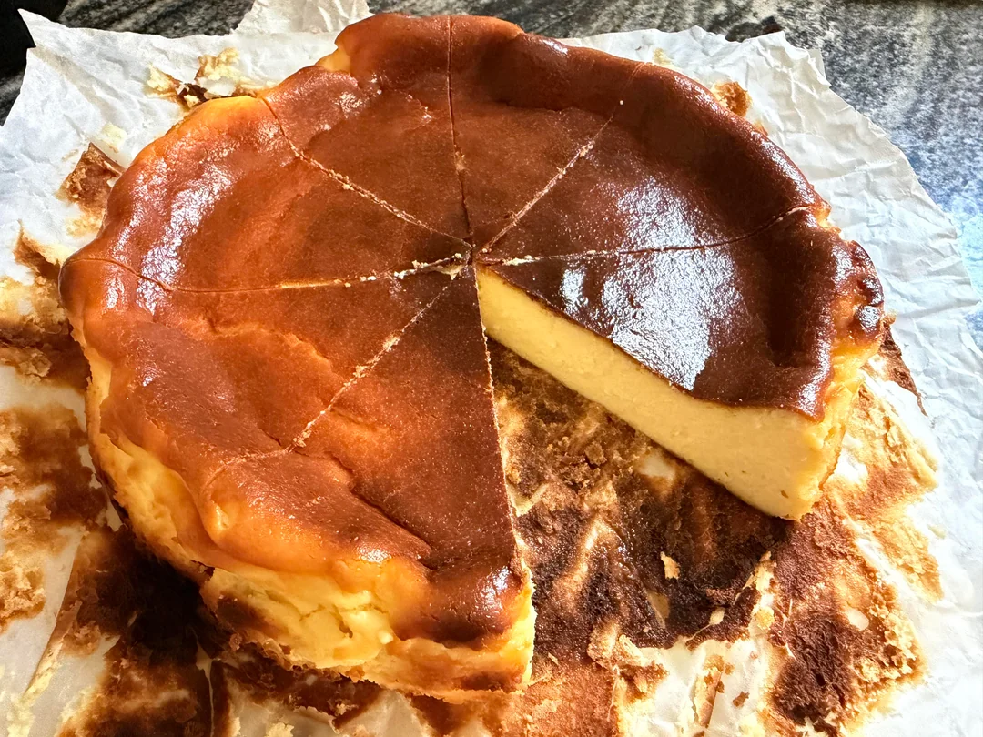 A Basque burnt cheesecake with a caramelized top, sliced into eight pieces, with one slice removed. The cheesecake sits on crumpled parchment paper atop a stone countertop.