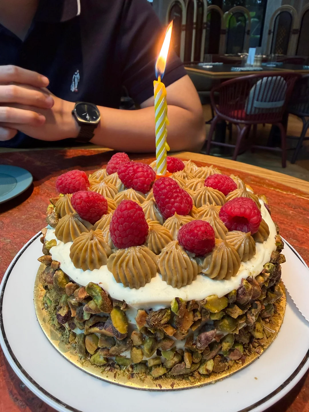 A cake topped with raspberries, frosting, and chopped nuts has a single lit candle. A person sits behind the cake with hands clasped, wearing a dark shirt and watch. The scene is set in a restaurant.