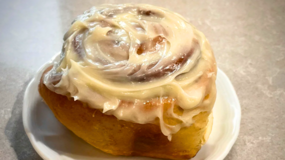 A large cinnamon roll topped with creamy white icing sits on a white plate, placed on a light-colored countertop, showcasing the irresistible charm of homemade baked goods.