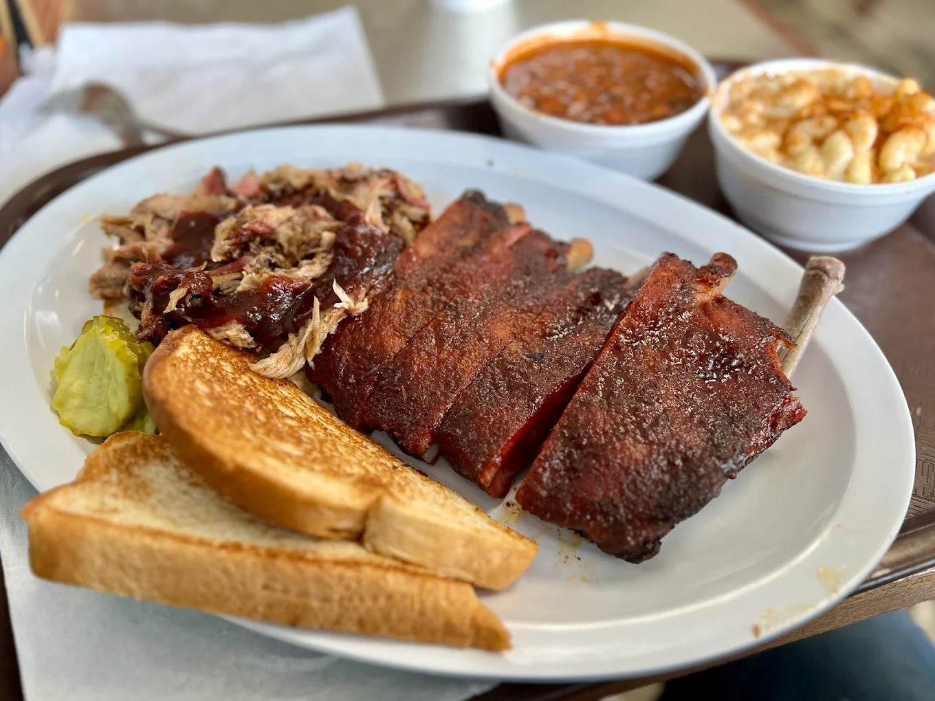 A plate with barbecue ribs, pulled pork, two slices of toasted bread, and pickles, served with sides of baked beans and macaroni and cheese in small bowls.