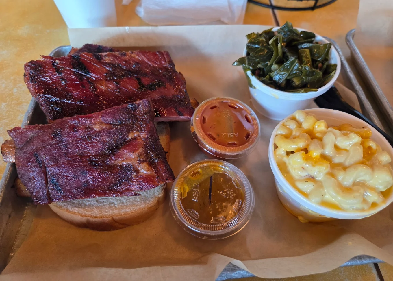 A tray of barbecue food features smoked ribs on slices of bread, small containers of barbecue sauce, a cup of macaroni and cheese, and a cup of cooked greens.