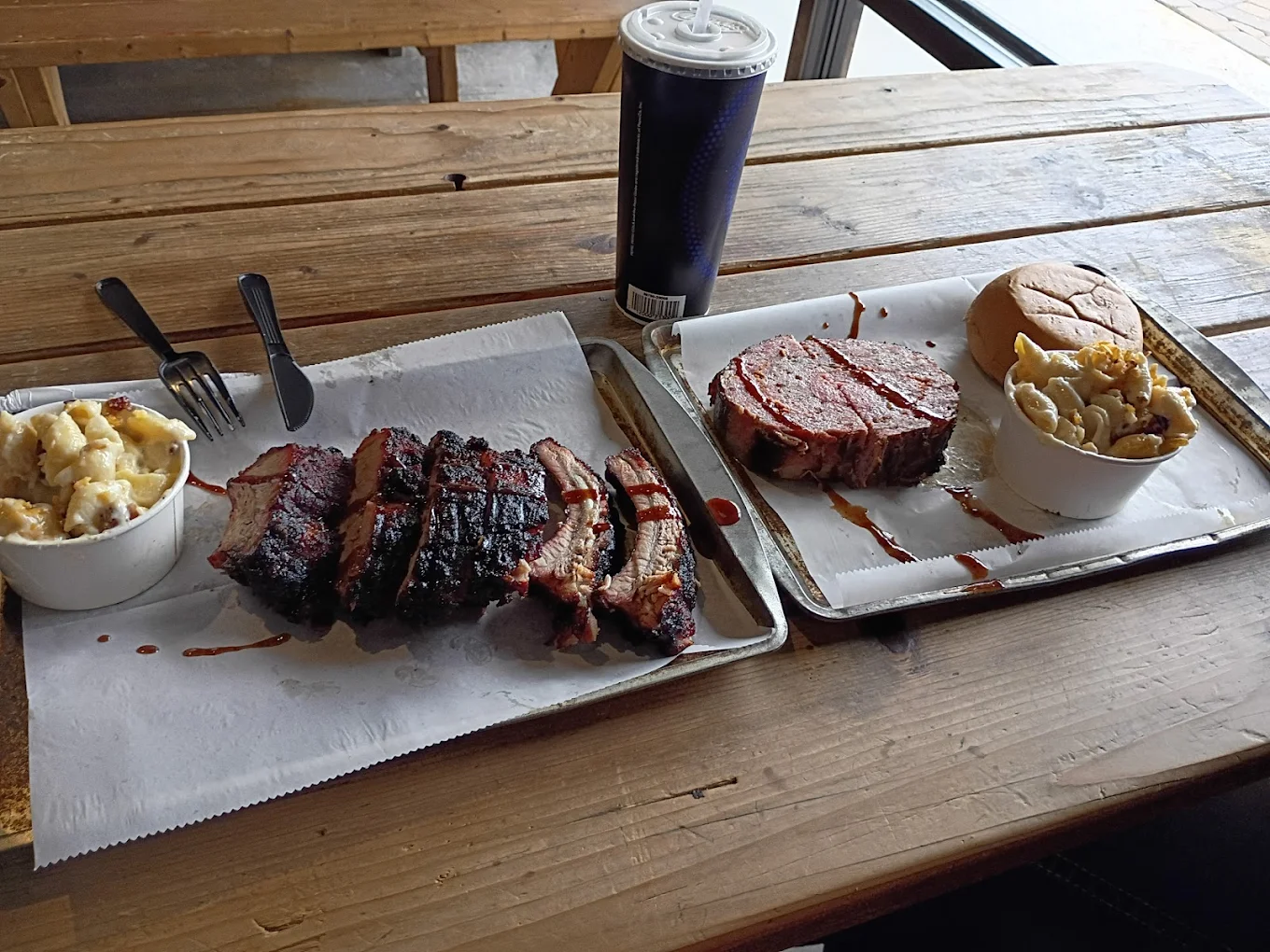 Two trays of barbecue food on a wooden table: one with sliced brisket and macaroni and cheese, the other with a thick slice of meat, a sandwich bun, macaroni and cheese, and a large drink cup in the background.