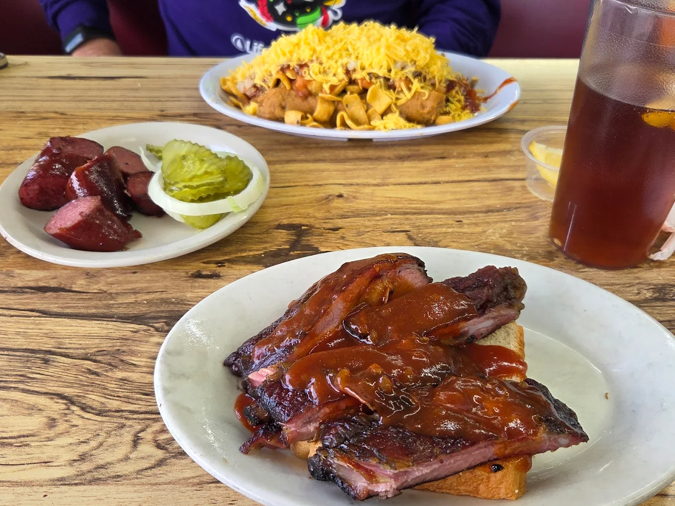 Three plates of barbecue food on a wooden table: sliced sausage with pickles, saucy ribs on toast, and a large plate of Frito pie with shredded cheese. An iced drink with lemon is beside the plates.
