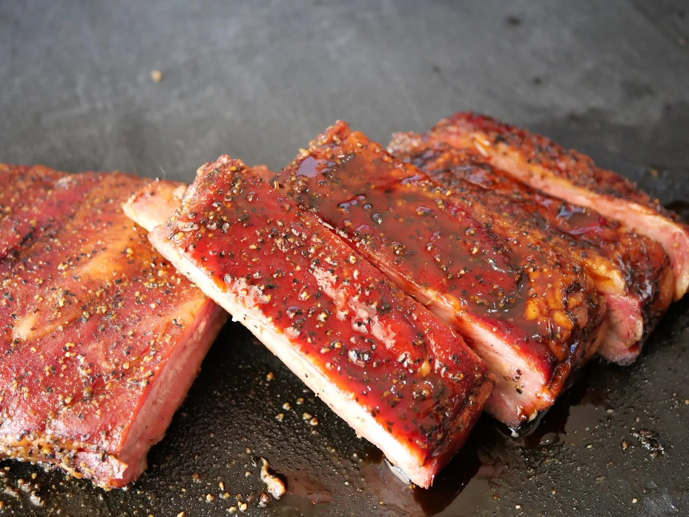 Three pieces of glazed, peppered barbecue ribs rest on a dark surface, glistening with sauce and juices. The meat appears tender with a rich, reddish-brown color and visible seasoning.