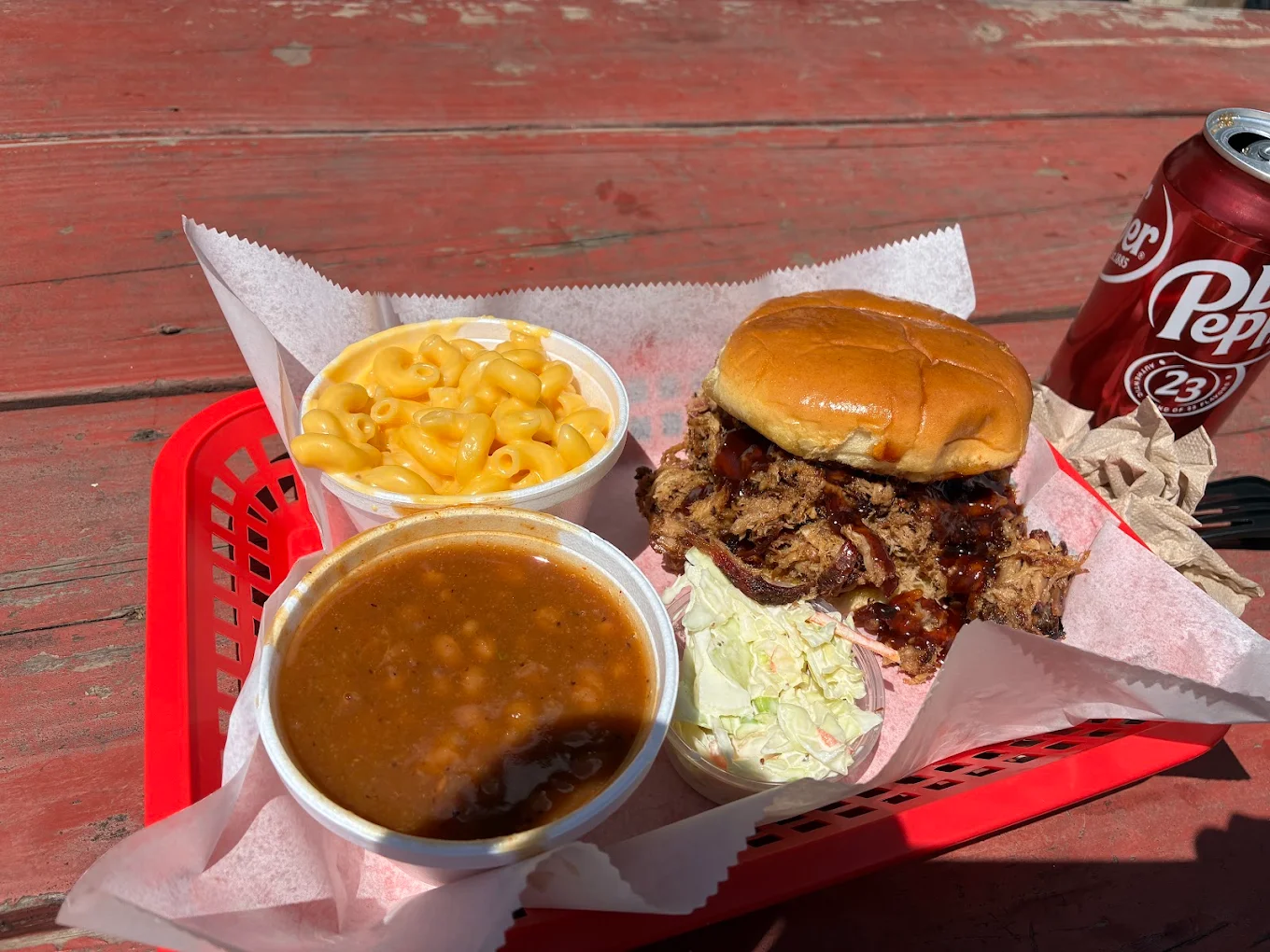 A red basket holds a pulled pork sandwich with barbecue sauce, a serving of coleslaw, baked beans, and macaroni and cheese. A can of Dr Pepper sits beside the basket on a weathered wooden table in bright sunlight.