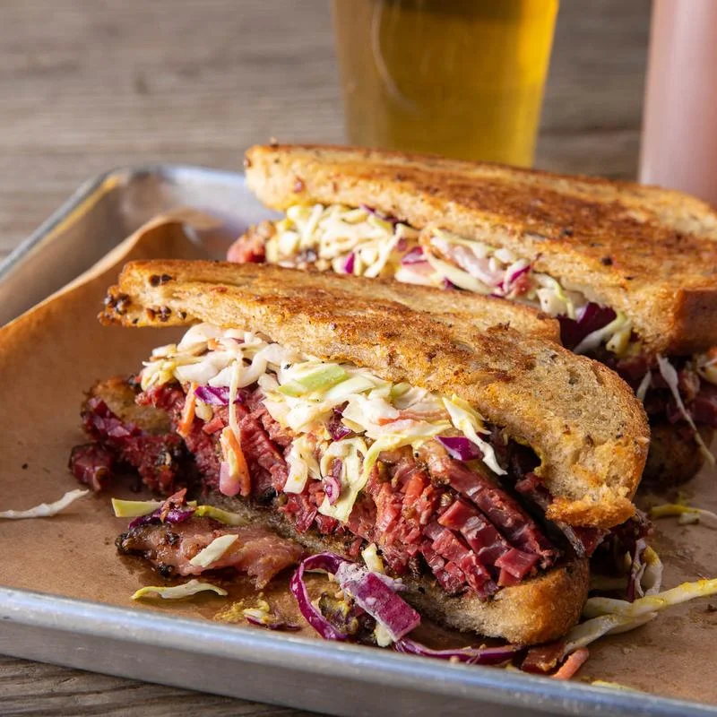A close-up of a sandwich with thick slices of pastrami, coleslaw, and grilled rye bread, served on a tray with a glass of beer and a bottle in the background.