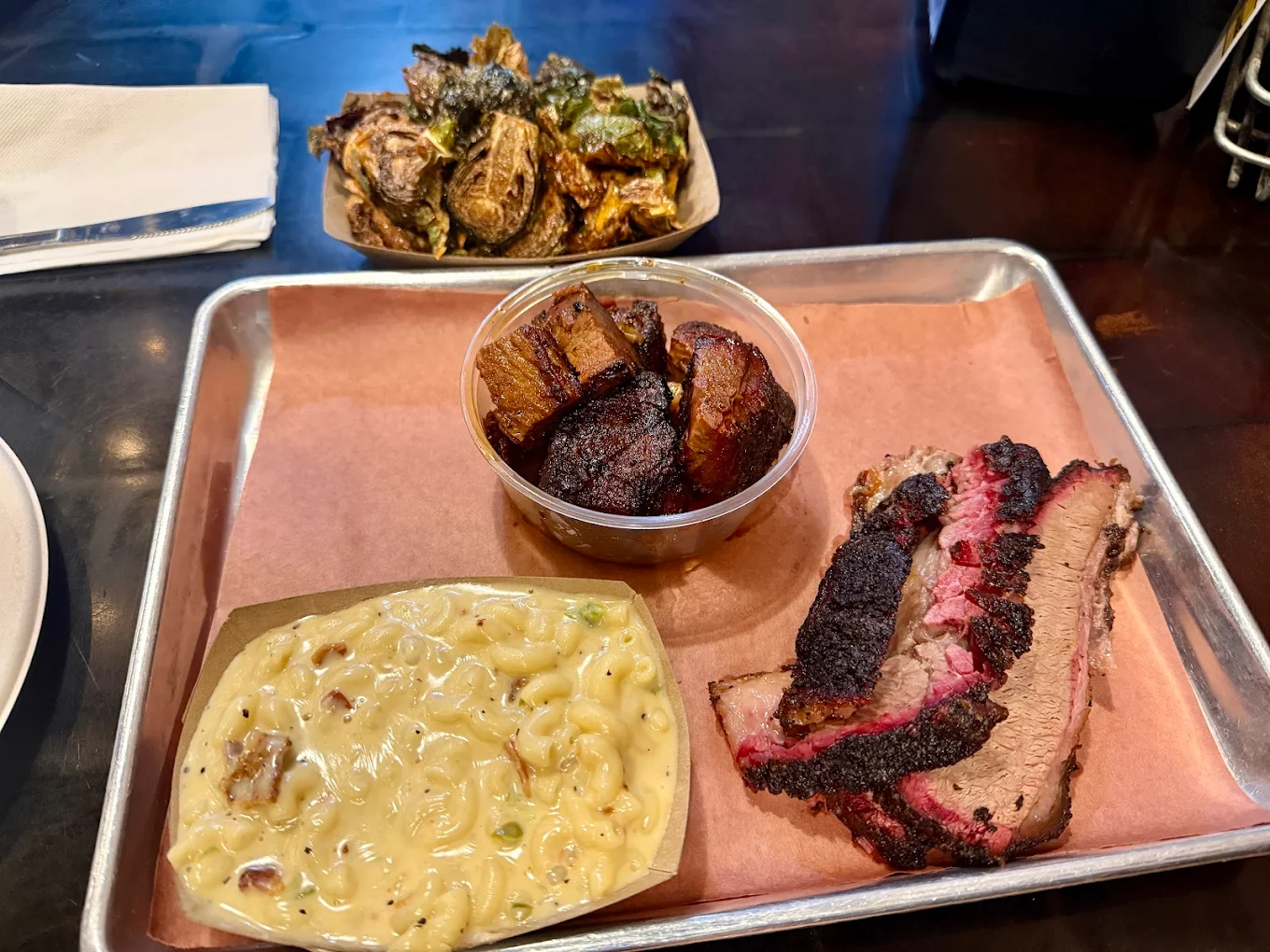 A metal tray holds macaroni and cheese, two slices of smoked brisket with a dark bark, and a cup of roasted pork belly; a separate plate in the background contains crispy Brussels sprouts.