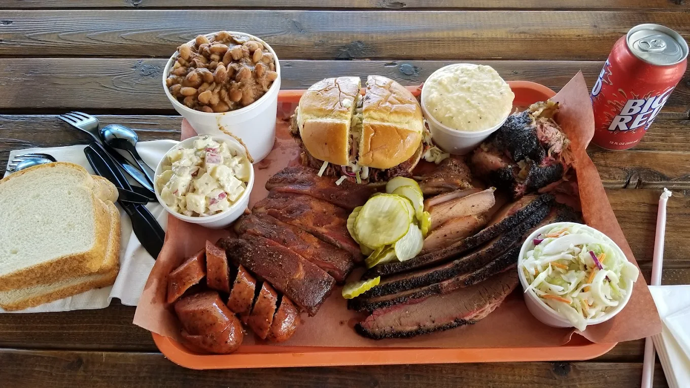 A tray of barbecue food with sliced brisket, sausage, ribs, pickles, a sandwich, potato salad, coleslaw, baked beans, white bread slices, and a can of Big Red on a wooden table.