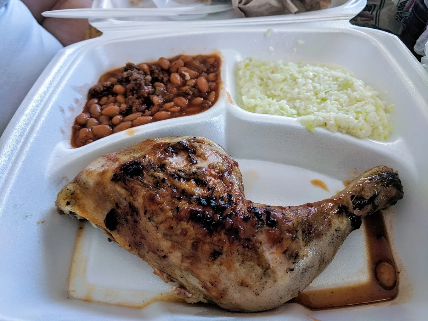 A divided foam takeout container with grilled chicken leg quarter, baked beans with ground meat, and a serving of coleslaw. Chopsticks and napkins are in the background.