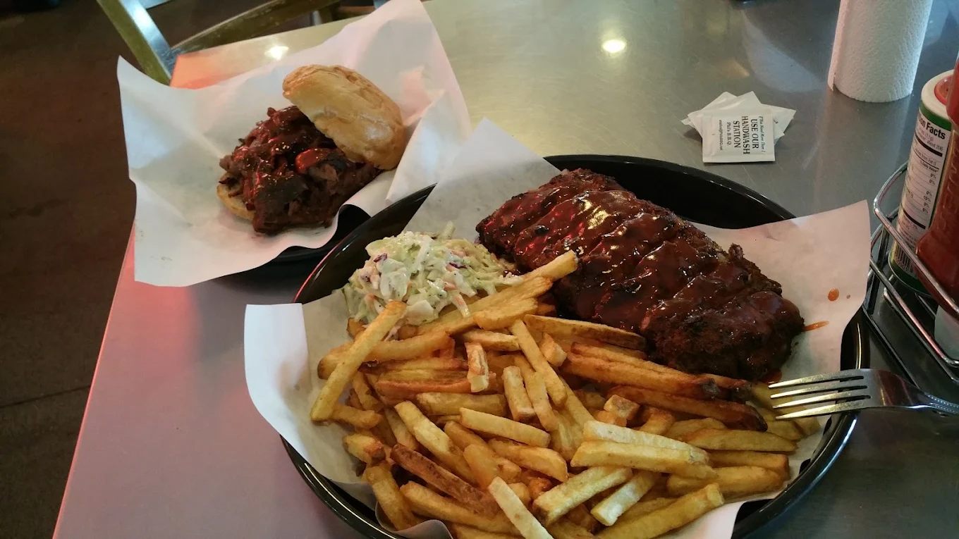 A plate of French fries, coleslaw, and barbecued ribs with sauce sits next to a sandwich filled with sliced meat on a bun, all on a metallic table. A fork and condiment packet are also visible.