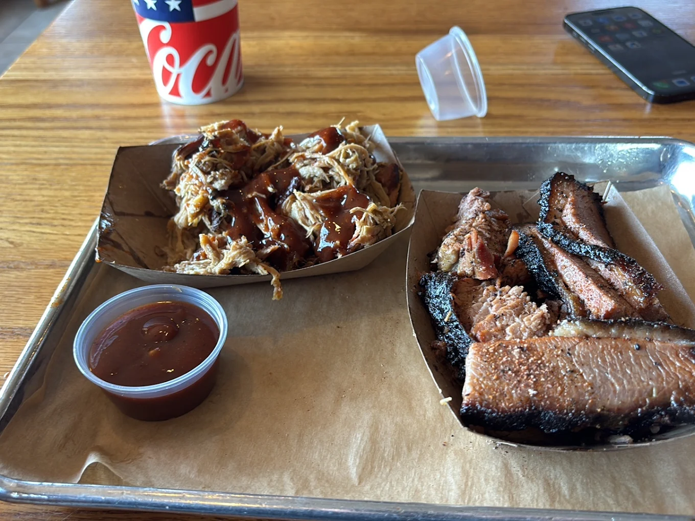 A tray with a serving of barbecued brisket slices, pulled pork topped with barbecue sauce, a small cup of extra sauce, a paper cup of soda, and a smartphone on a wooden table.