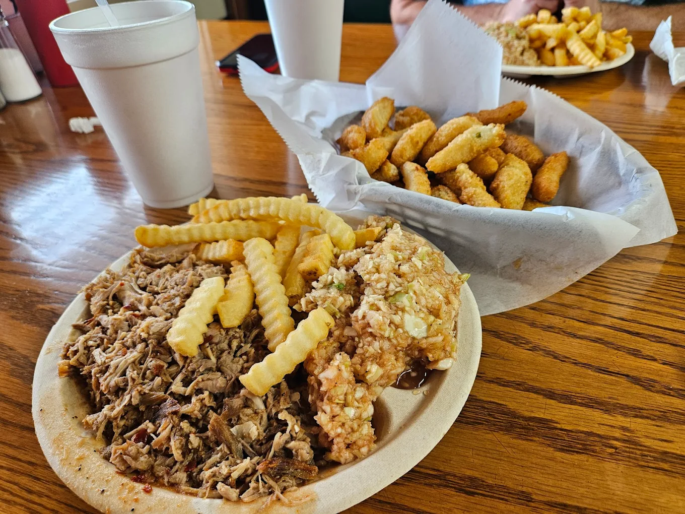 A plate of pulled pork, crinkle-cut fries, and coleslaw sits on a wooden table next to a basket of fried okra and two white foam cups. Another plate of fries is partially visible in the background.