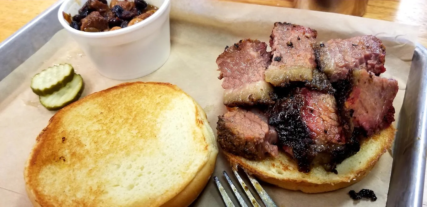 A tray with an open sandwich: slices of smoked brisket on the bottom half of a bun, the top bun to the side, a few pickle slices, a fork, and a small cup of baked beans in the background.