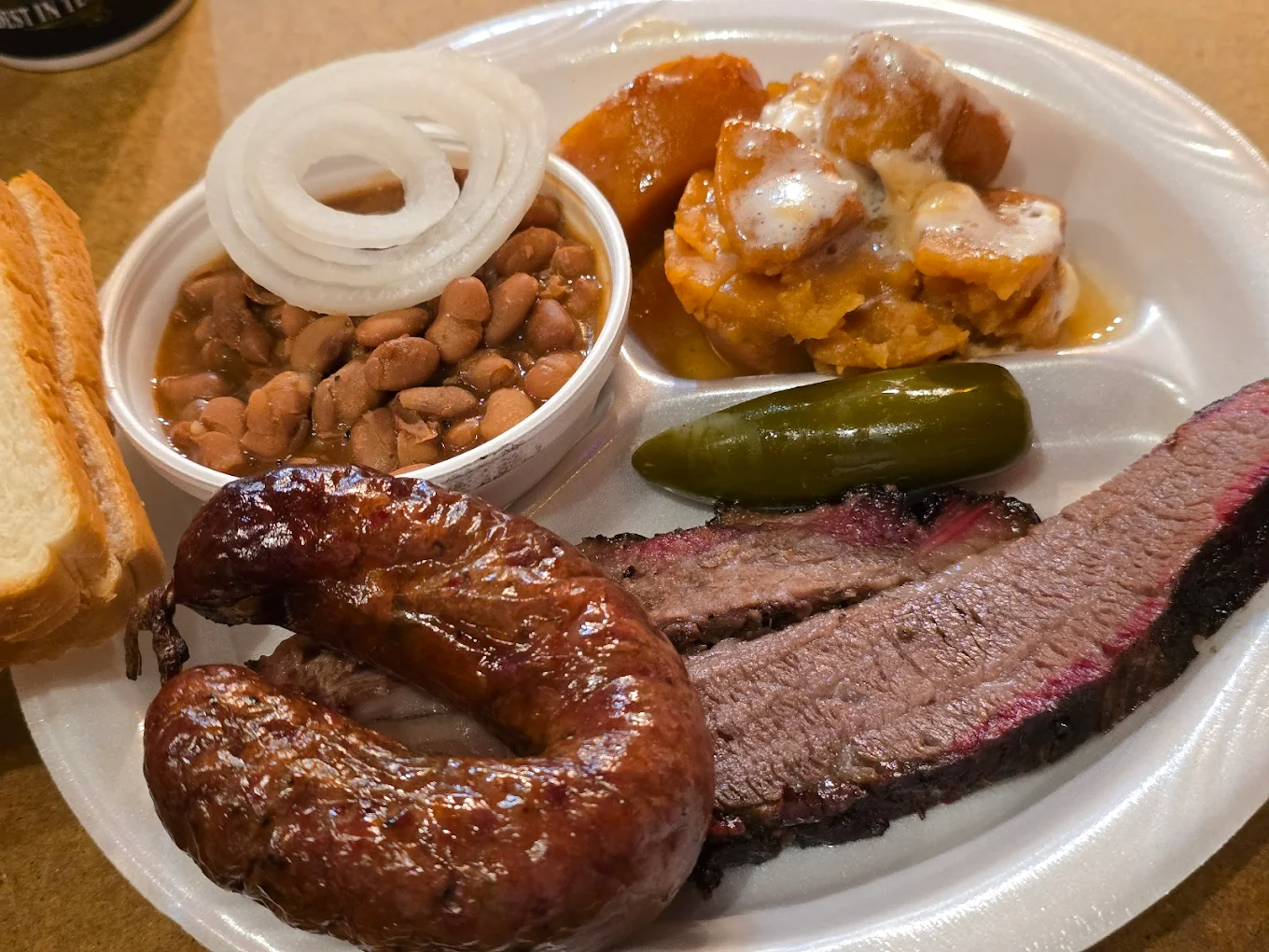 A plate of barbecue food featuring sliced brisket, a sausage link, pickles, pinto beans, sweet potatoes with sauce, raw onion slices, and a slice of white bread on a white foam plate.