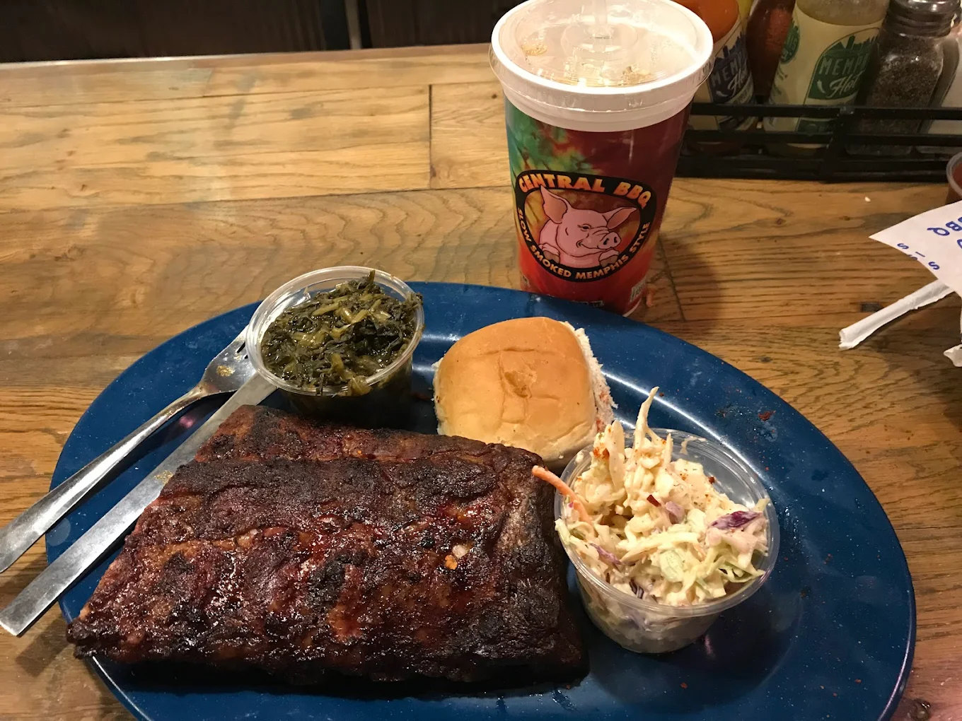 A blue plate with a serving of barbecue ribs, a bread roll, coleslaw, and cooked greens, next to a drink cup labeled "Central BBQ" on a wooden table.
