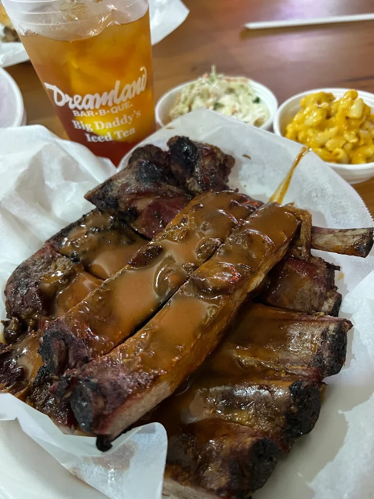 A plate of saucy barbecue ribs sits on parchment paper, with bowls of coleslaw and macaroni and cheese in the background, and a large cup of iced tea labeled "Dreamland Bar-B-Que Big Daddy's Iced Tea.