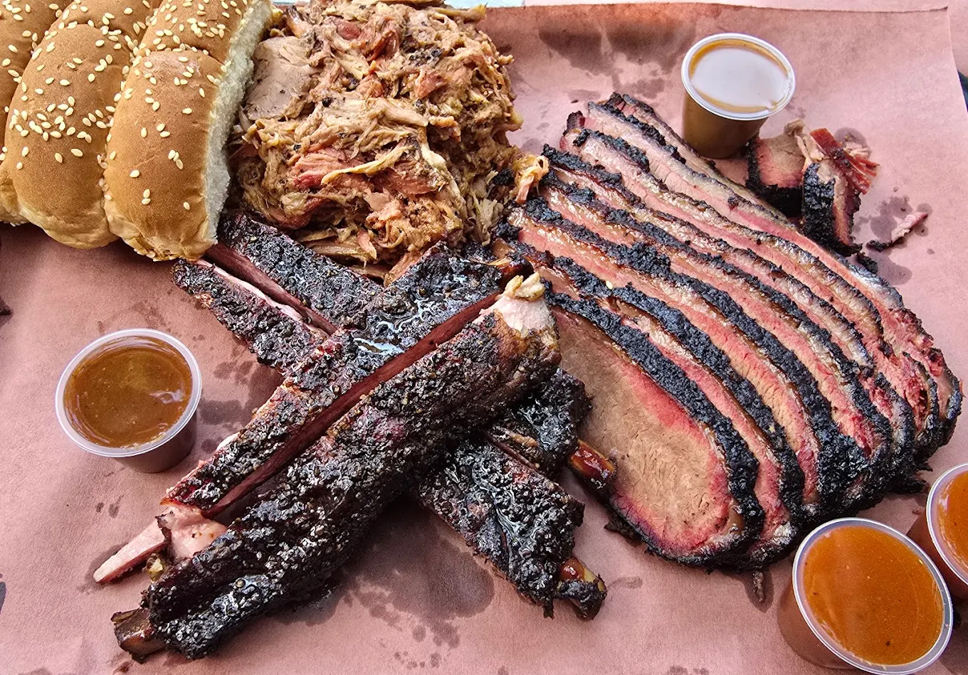 A tray of barbecue food with sliced brisket, ribs, pulled pork, three sesame seed buns, and several small cups of barbecue sauce on brown butcher paper.