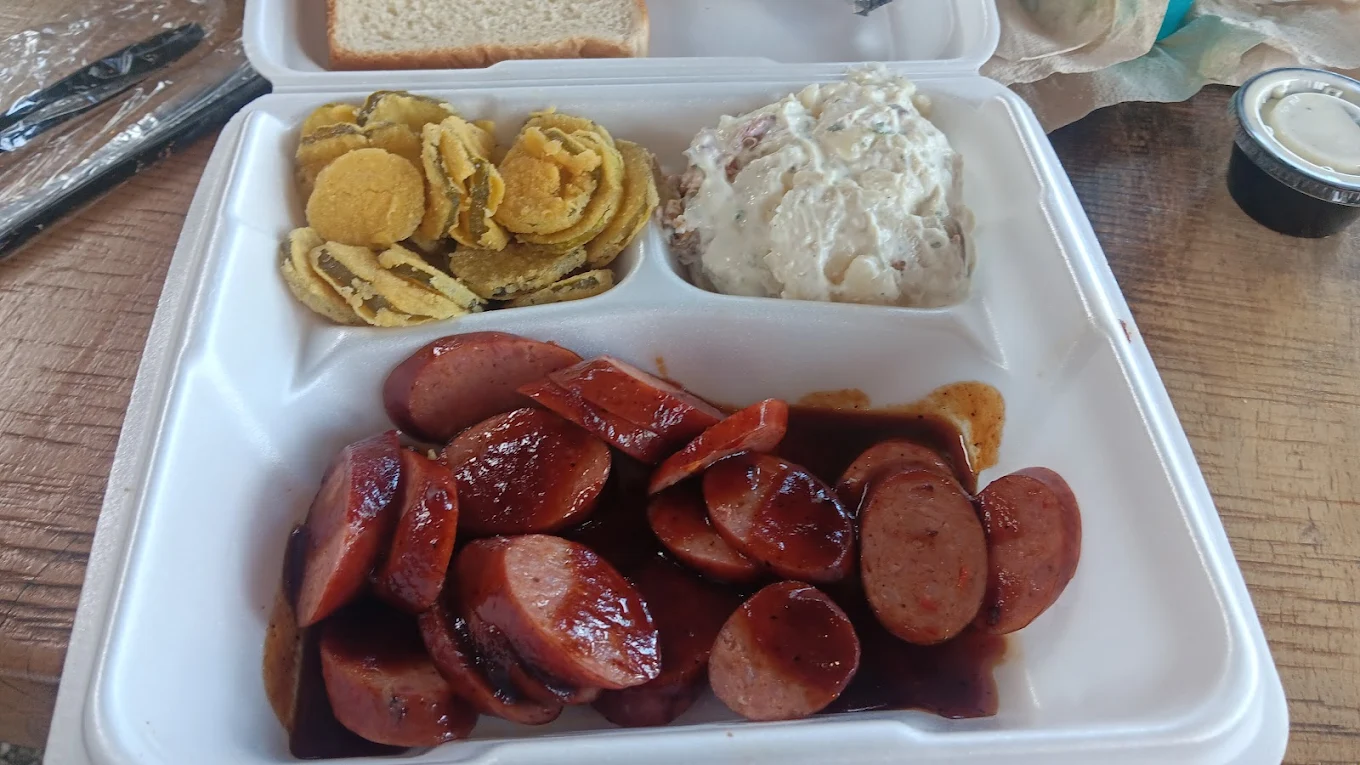 A takeout container with sliced sausage in barbecue sauce, fried pickles, potato salad, a slice of white bread, and a cup of ranch dressing on a wooden table.