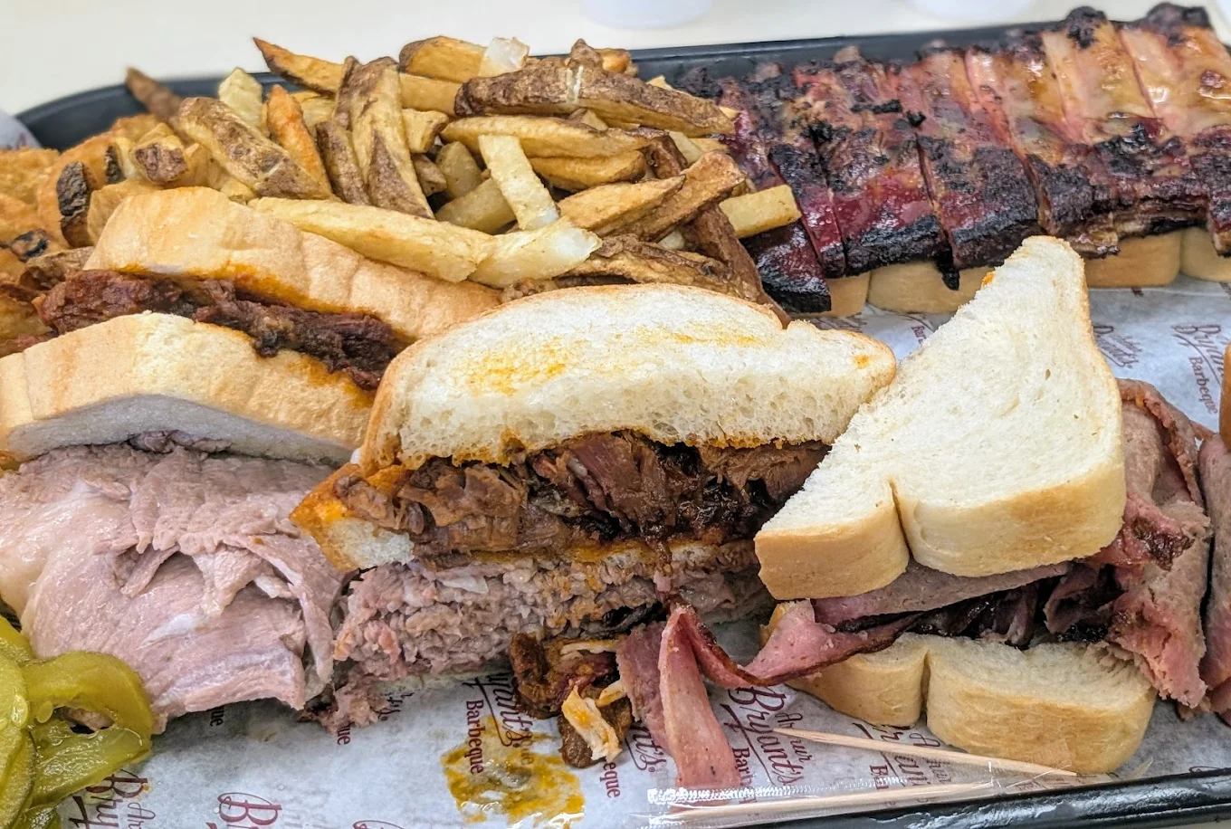 A tray filled with barbecue food, including sliced white bread sandwiches stuffed with various smoked meats, a pile of French fries, pickled jalapeños, and a rack of glazed ribs in the background.