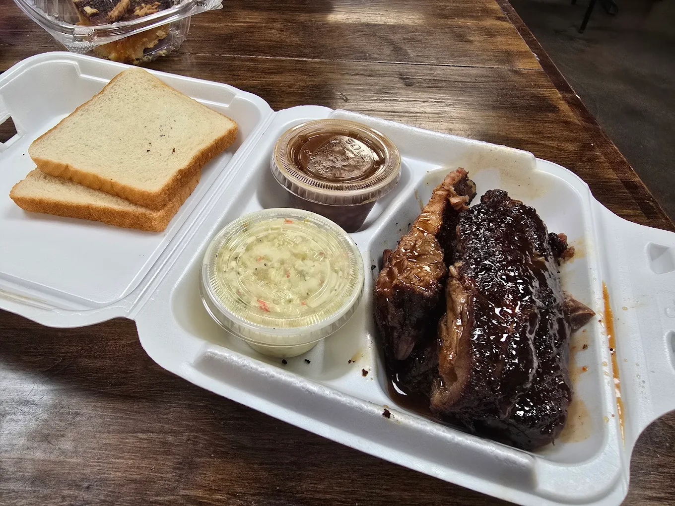 A takeout container with two slices of white bread, a serving of saucy barbecue beef brisket, a cup of coleslaw, and a cup of barbecue sauce on a wooden table.