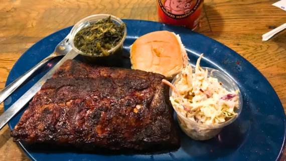 A blue plate with a serving of barbecued ribs, a bun, leafy greens in a plastic cup, and coleslaw; a drink with a pig logo sits in the background on a wooden table&mdash;a feast often found at cheap BBQ restaurants.