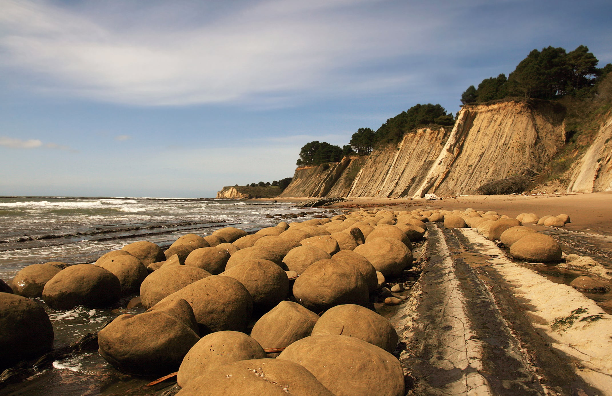 Large round boulders scattered along a sandy beach with steep, tree-topped cliffs in the background under a partly cloudy sky. Waves are gently approaching the shore.