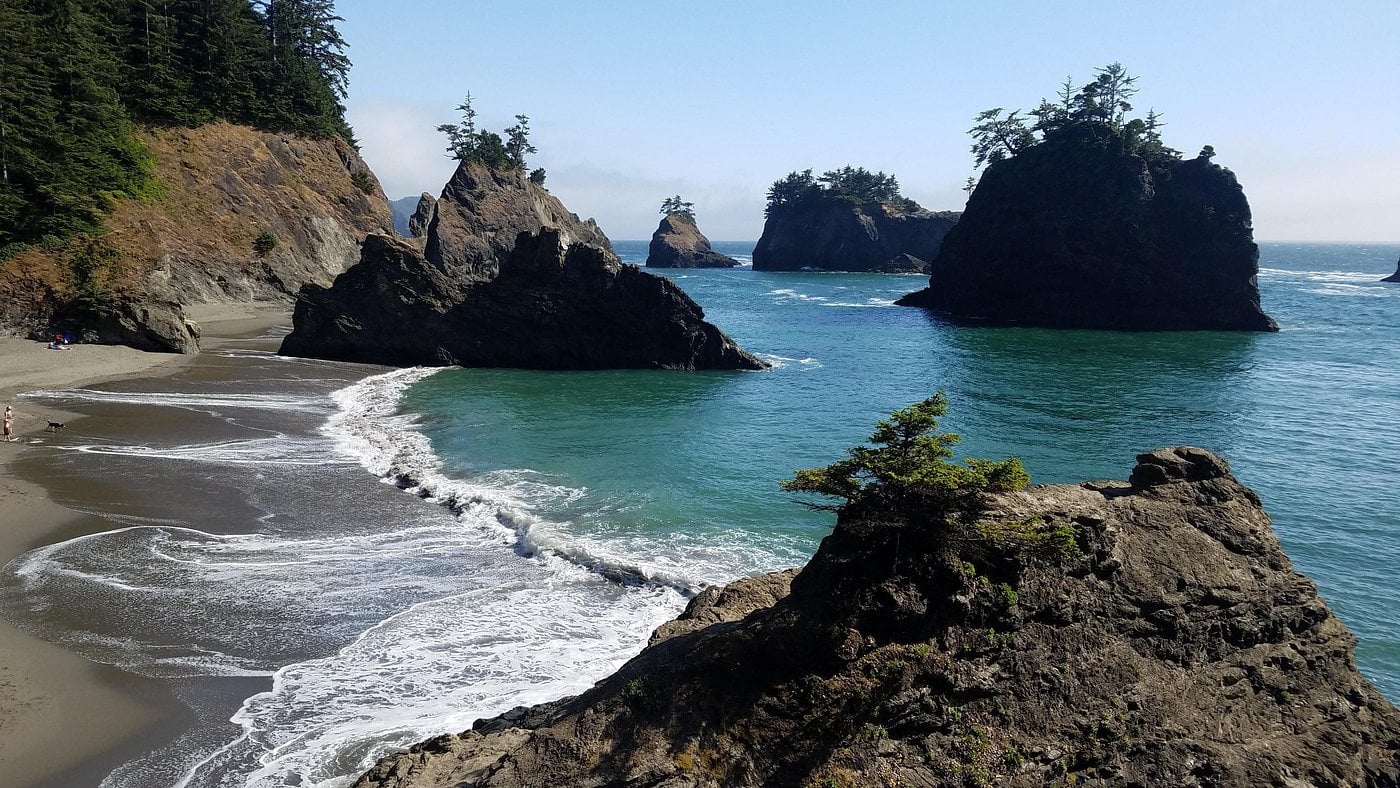 Rocky sea stacks and lush evergreen trees line a curving sandy beach beside turquoise waves under a clear blue sky, with forested cliffs in the background.