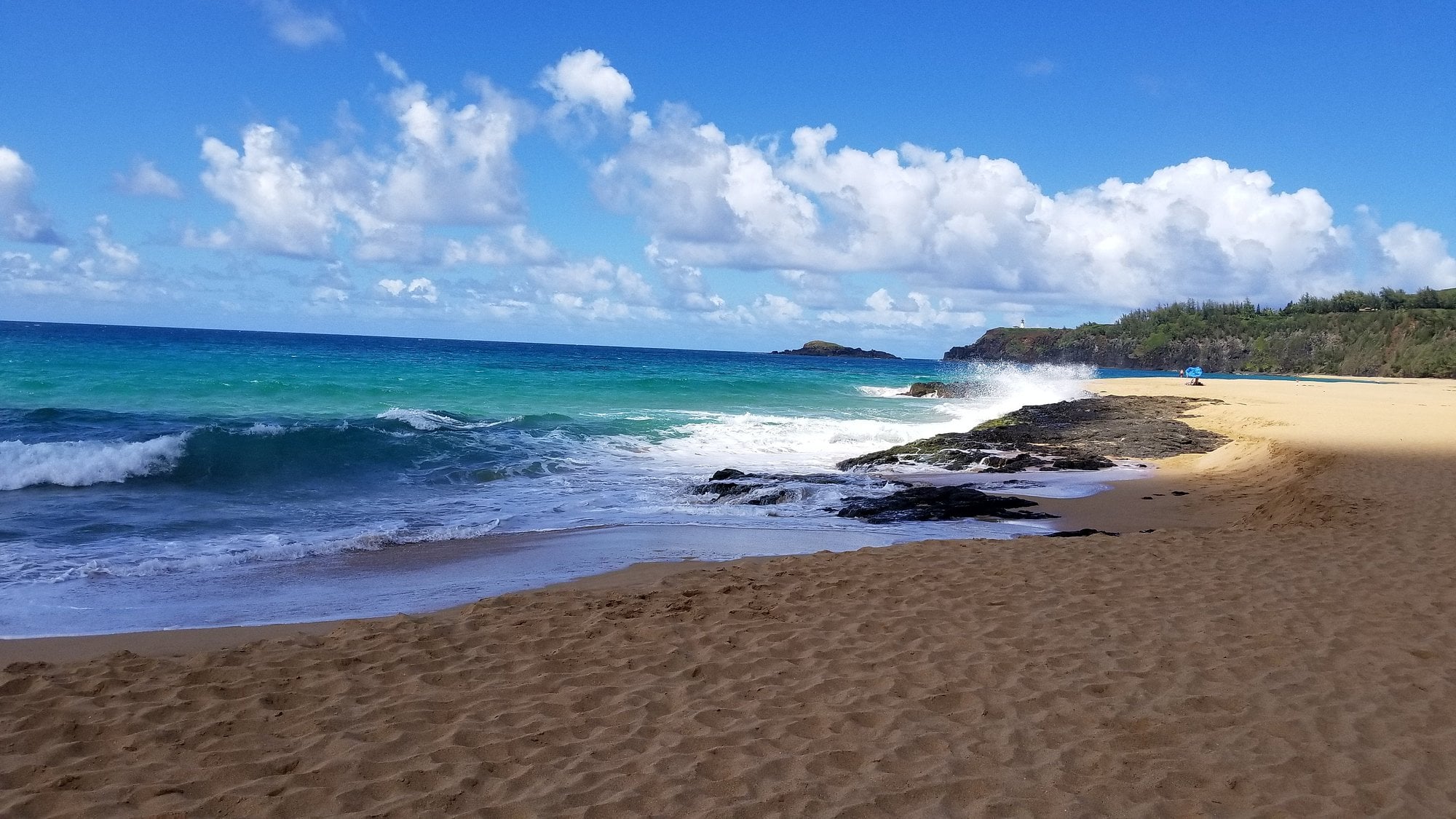 Sandy beach with turquoise waves crashing on rocks, under a bright blue sky with white clouds; distant greenery lines the horizon.