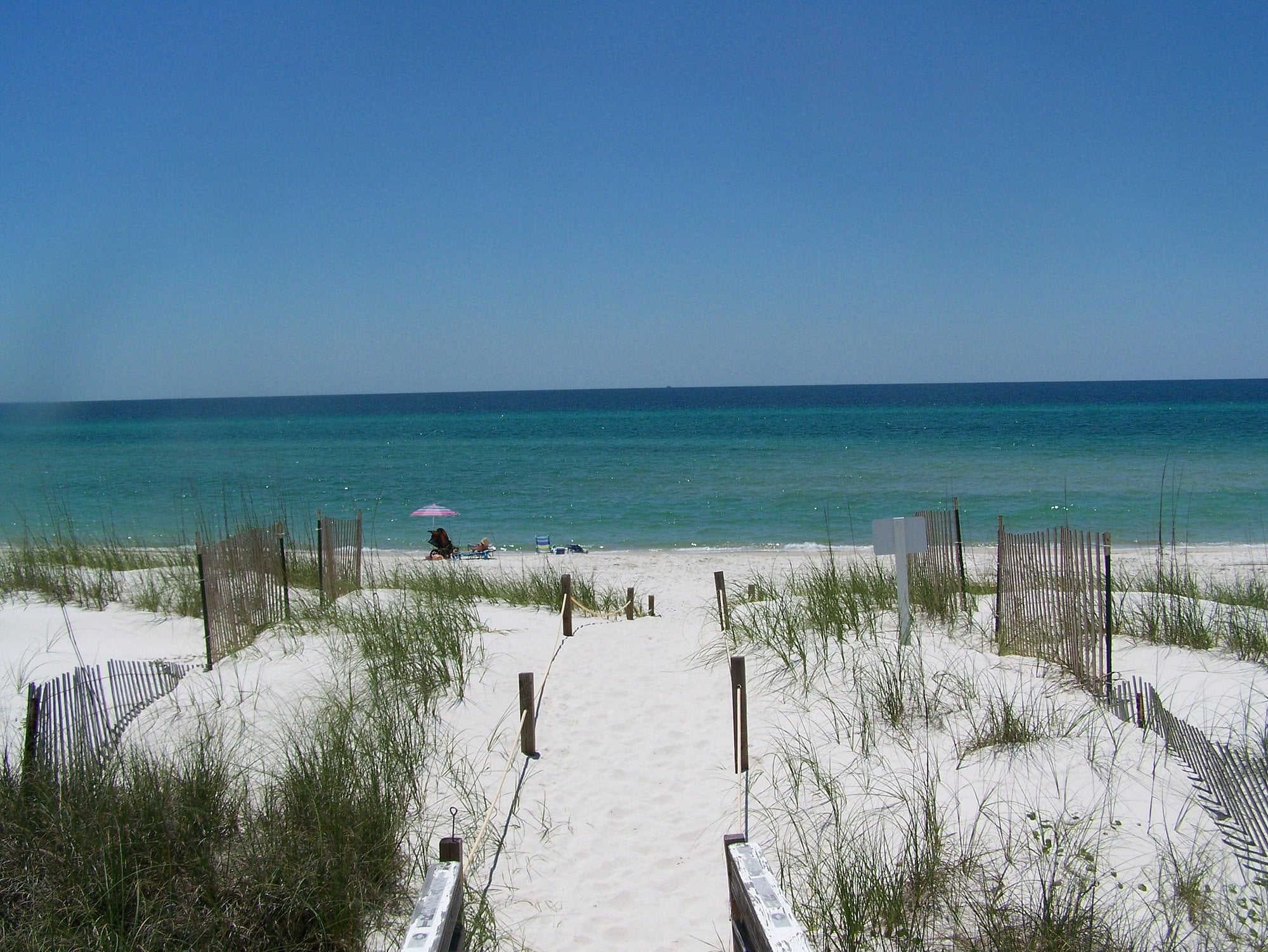 A sandy path lined with grasses and wooden fences leads to a beach with white sand and turquoise water under a clear blue sky. A few people and a pink umbrella are visible near the shoreline.