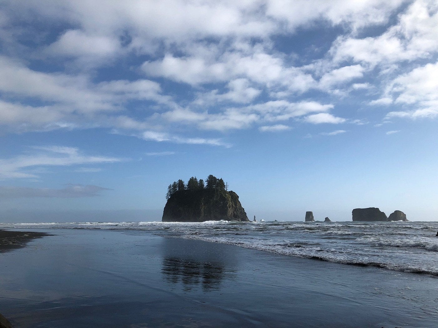 A large rock island with trees stands in the ocean near the shore under a partly cloudy sky, with smaller sea stacks in the distance and gentle waves meeting the sandy beach.