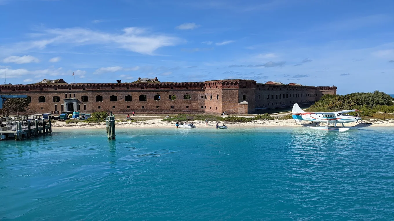 A historic brick fort stands on a sandy beach beside turquoise water, with a few small boats and a seaplane docked along the shore under a blue sky with scattered clouds.