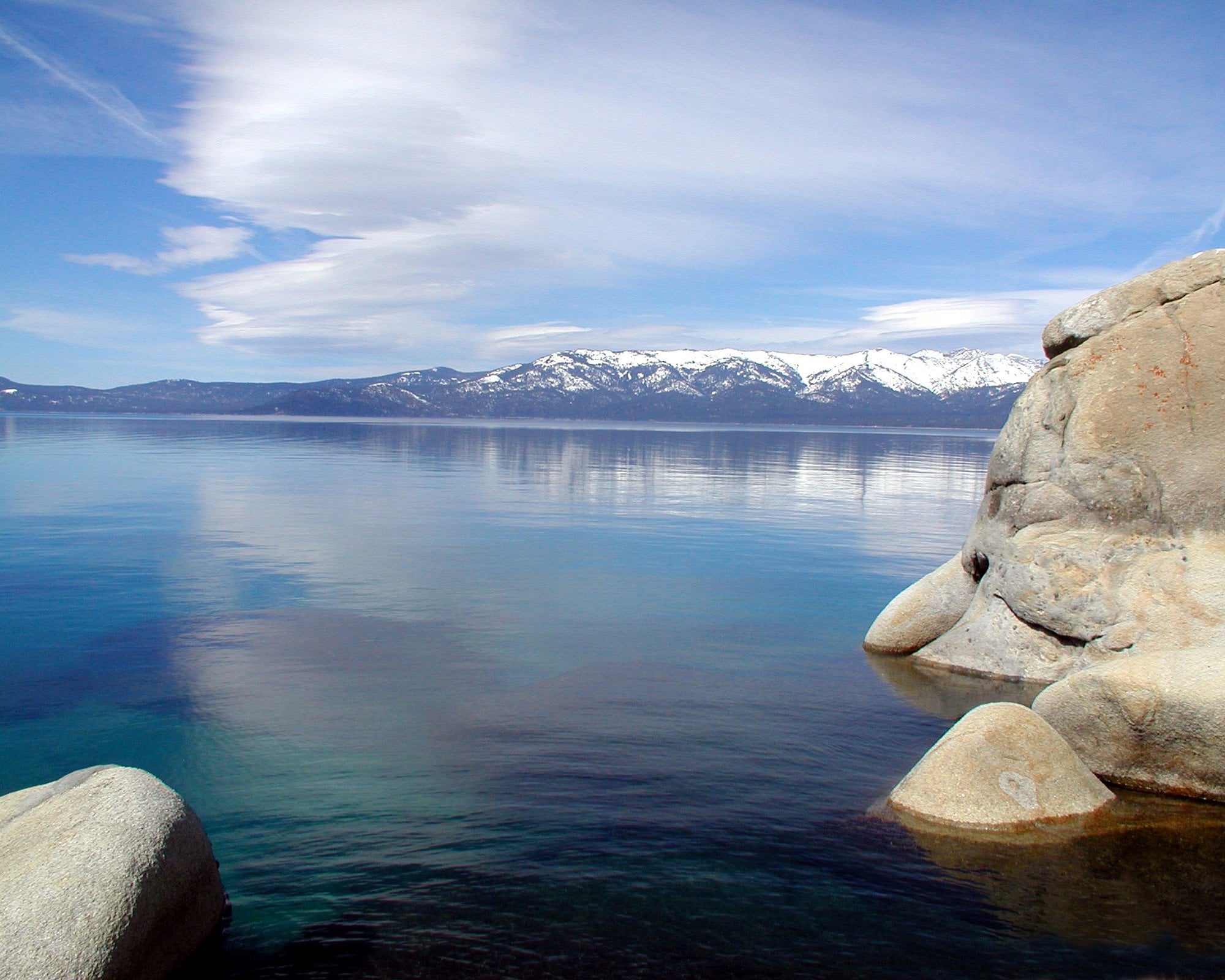 Clear lake with large boulders in the foreground, reflecting the sky and clouds. Snow-capped mountains are visible in the distance under a blue sky with wispy clouds.