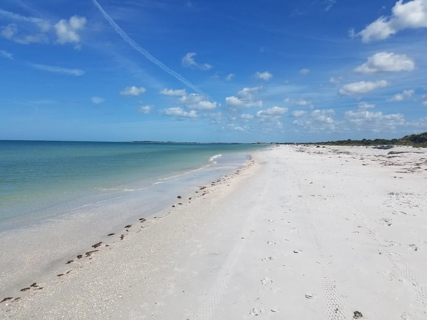 A pristine, empty beach with white sand, gentle waves, and clear blue water stretches into the distance under a bright blue sky with scattered clouds. Tire tracks and seaweed line the shore.